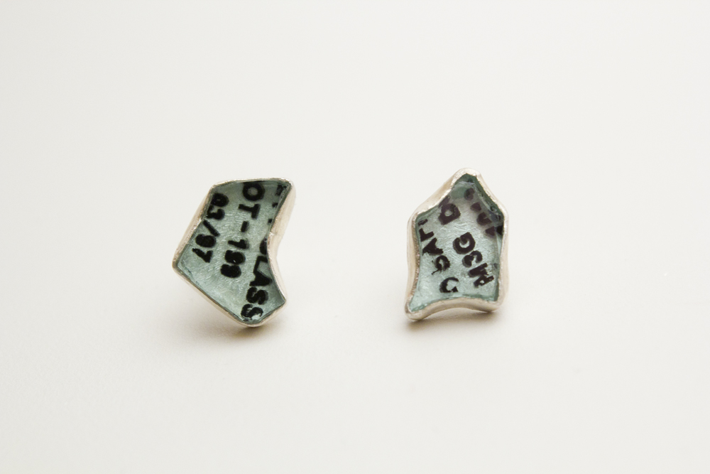 A pair of sterling silver stud earrings set with freeform chunks of green-tinted car glass with fragments of text, set in sterling silver bezels.