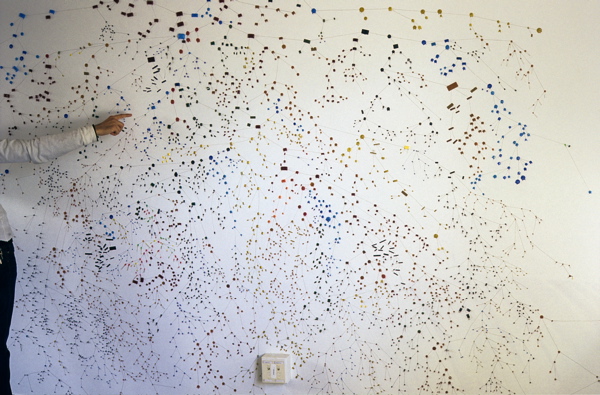 10 foot stretch of white wall covered in tiny multicoloured dots connected by lines with a man's hand pointing to the wall for scale.