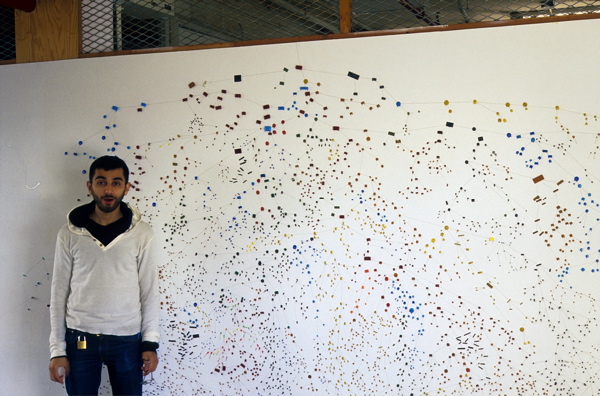 10 foot stretch of white wall covered in tiny multicoloured dots connected by lines with a man standing in front of the wall for scale.